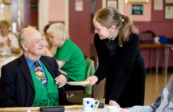 Female clergy member serving plate of food to older gentleman at table 