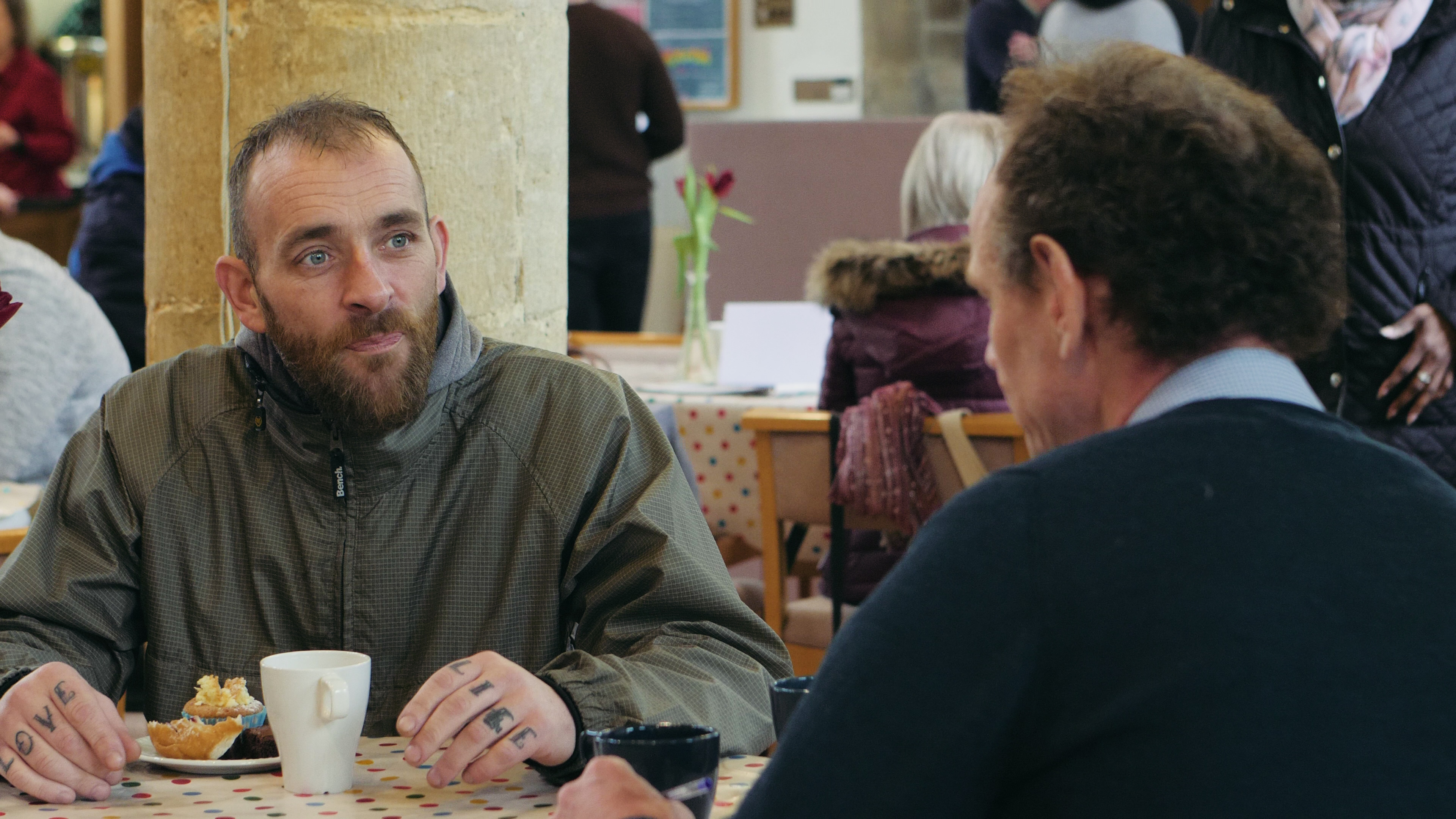 A man sits in a church drinking tea and eating cake while chatting to another man.
