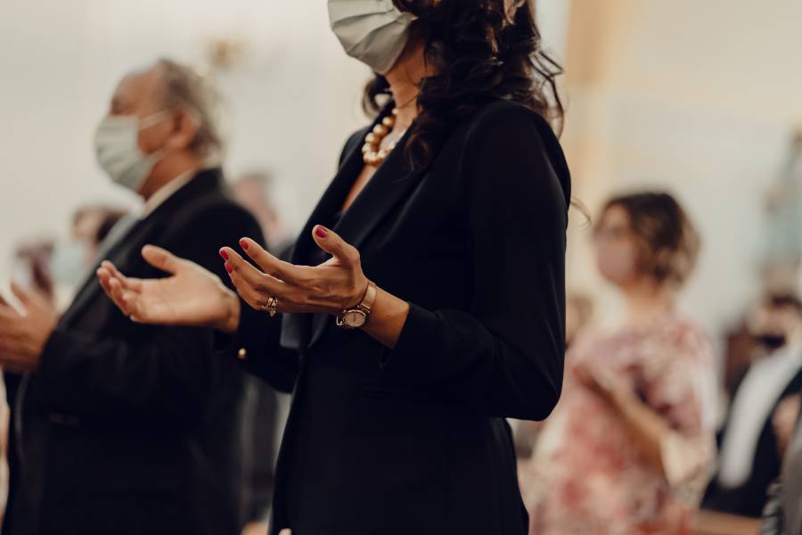A woman is shown with outreached hands in prayer wearing a facemask in church 