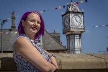 Woman with purple hair leaning against a wall and smiling in Downham Market with a clock tower in the background