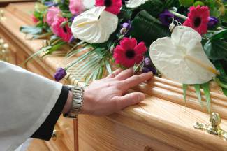 Close up of a vicar's hand touching a coffin lid. Flower display also on the lid.