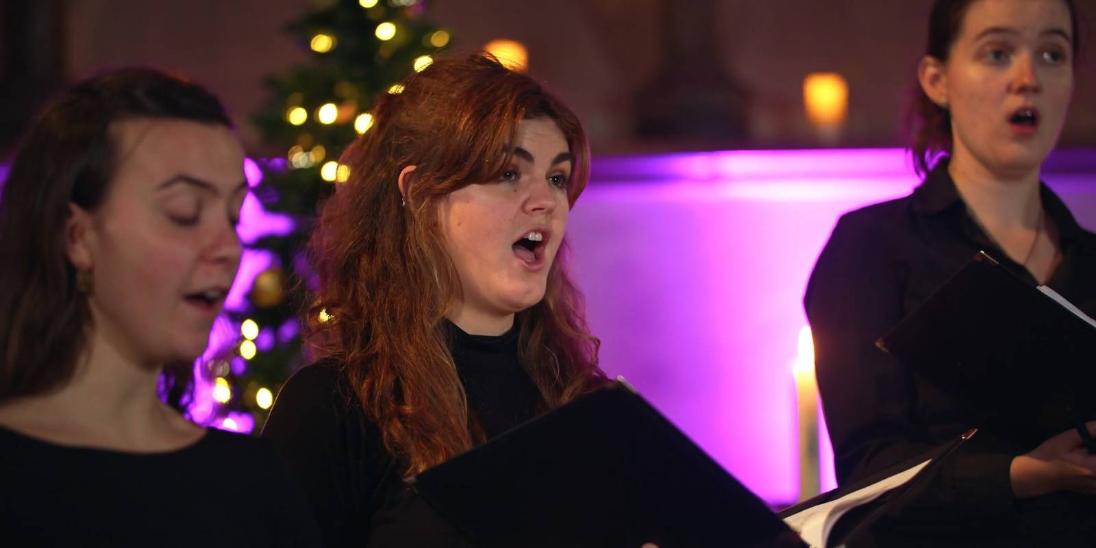 Three members of St Martin's Voices singing with a Christmas tree in soft focus
