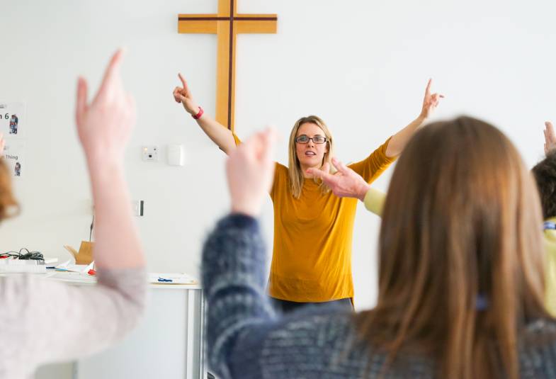 Woman with hands in the air leading worship, cross on wall behind 