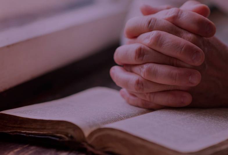 Clasped hands praying over an open Bible with a purple tint