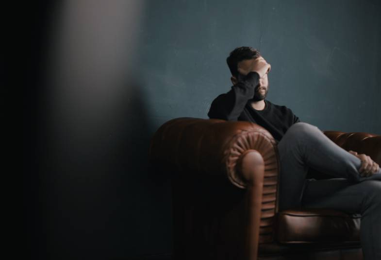 A man holds his head while sitting on a sofa looking sad 