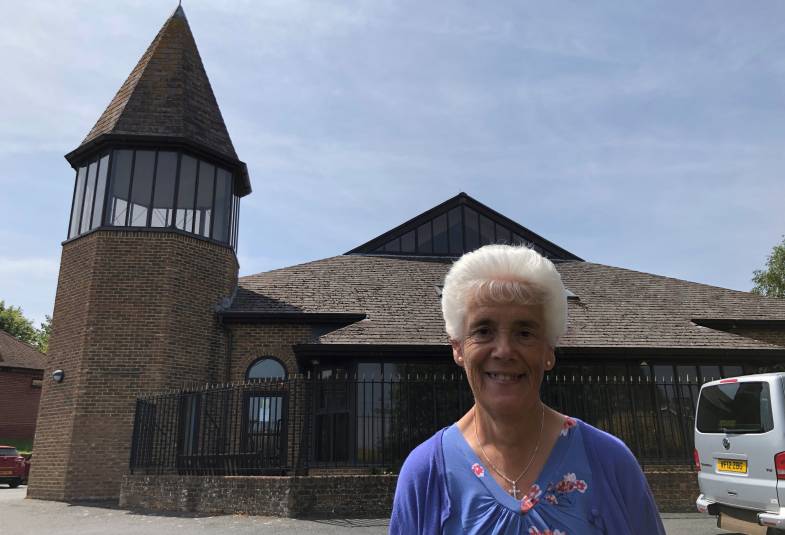 Sue is shown standing outside her church which is behind her she is smiling wearing a blue top