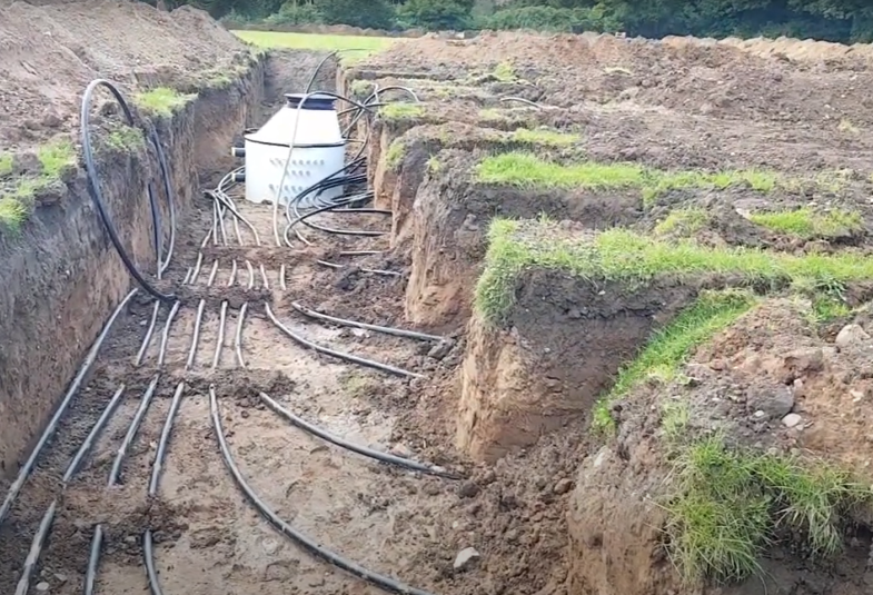 A heat pump is shown in a trench with pipes emerging from it and running along the muddy trench floor