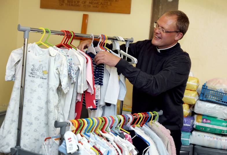 A vicar is putting baby clothes on a clothes rack and smiling