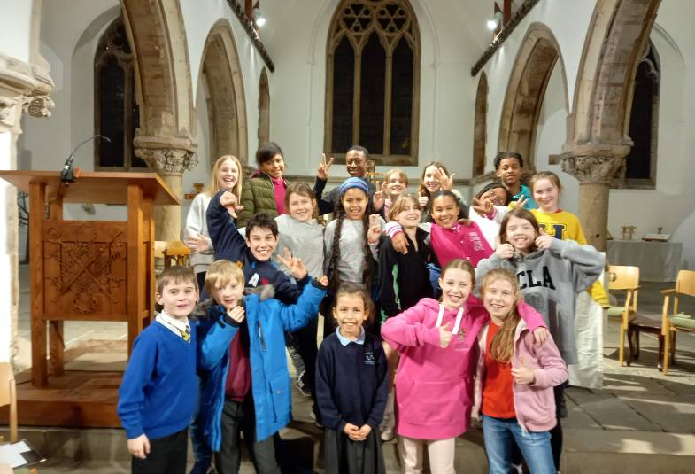 A diverse group of children and young people gathered for a photo in a church, all smiling and using thumbs up hand gestures