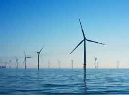 Wind turbines are shown off-shore in the sea with a ship in the background