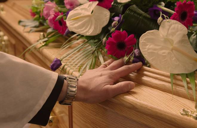 Clergy laying hand on coffin, flower arrangement on top 