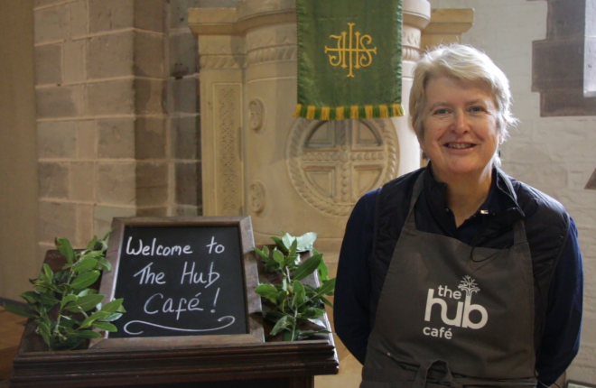 Woman wearing branded apron stands next to welcome sign at church cafe