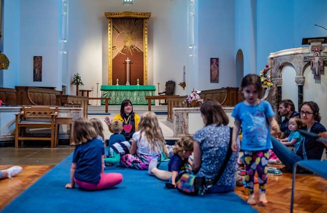 Vicar telling children a story. They are sitting on the blue carpeted floor of the main aisle of the church.