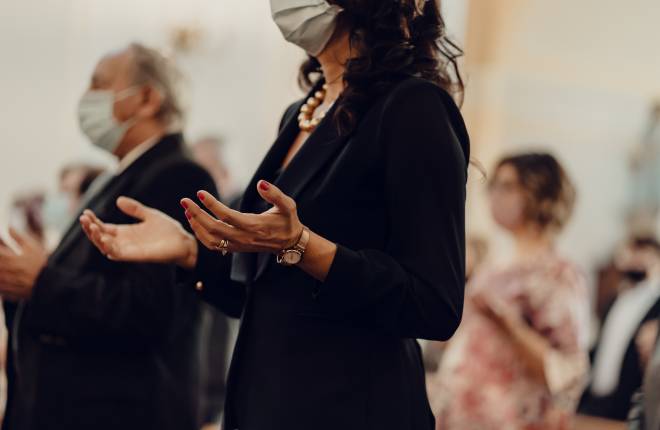 A woman is shown with outreached hands in prayer wearing a facemask in church 