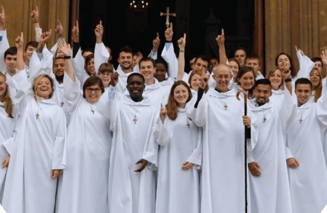 Members of the Community of St Anselm standing outside Lambeth Palace in their white robes, smiling at the camera and pointing their index finger in the air.