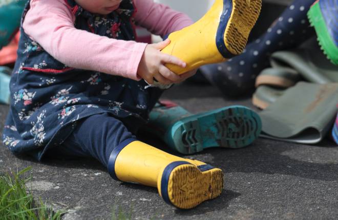 A little girl toddler is sitting on the ground pulling on yellow wellington boots