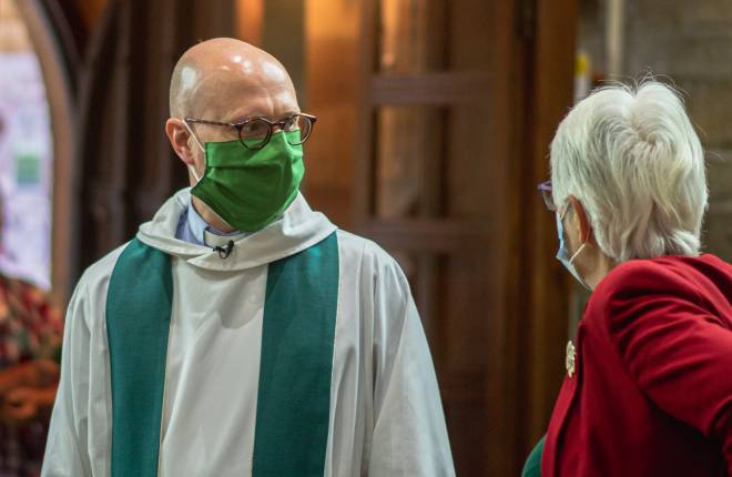 A vicar with a green mask on is seen smiling inside a church to a parishoner